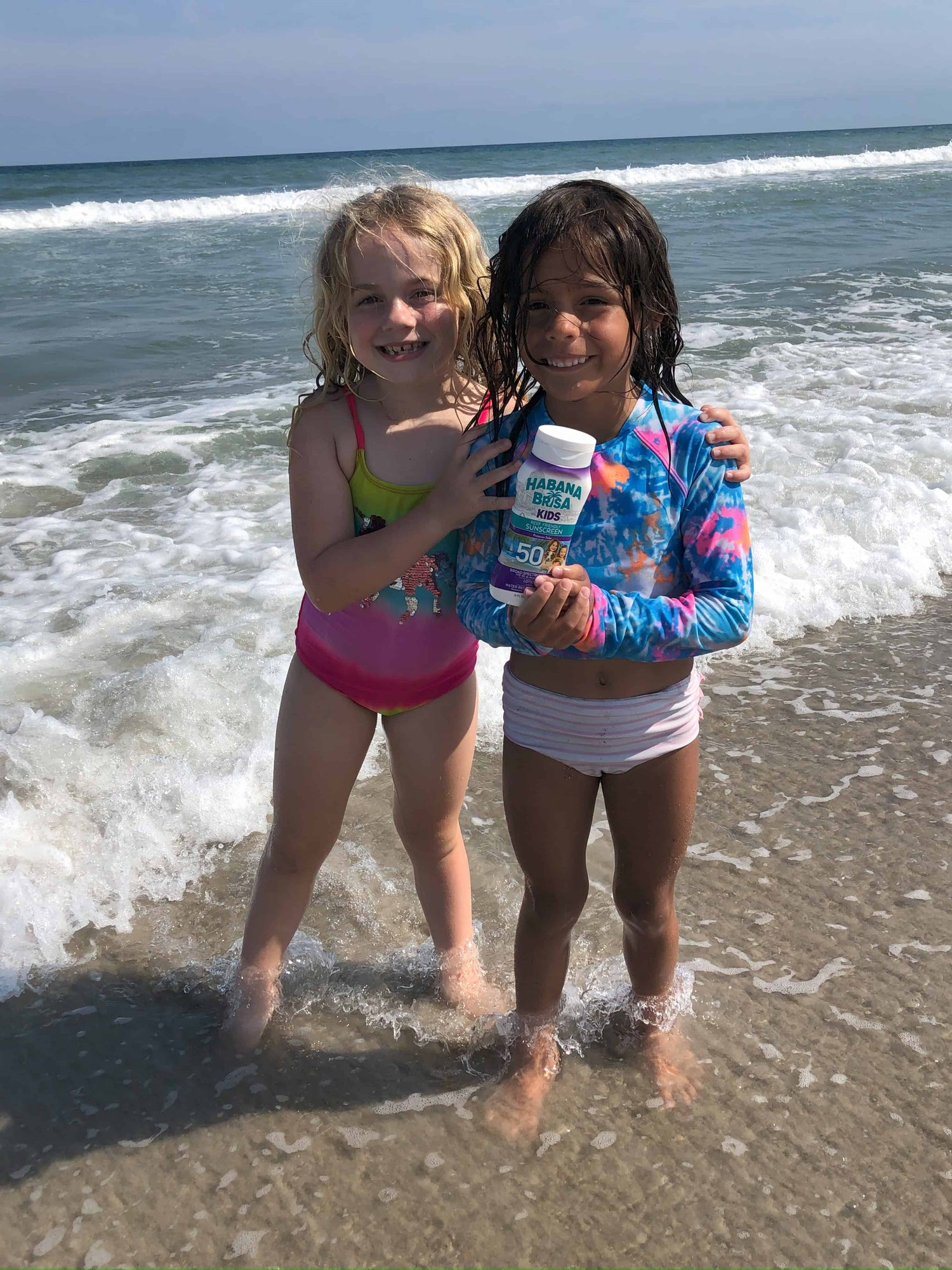 Two smiling girls at the beach holding Habana Brisa Kids SPF 50 sunscreen, enjoying sun protection with reef-safe, water-resistant sunscreen for kids.